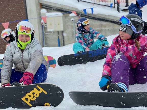 Dzieci siedzą na śniegu z załoąonymi deskami snowboardowymi. 